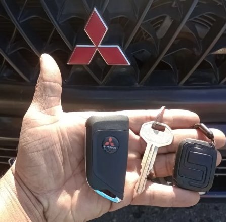 Mão do chaveiro da DK segurando uma chave canivete, uma chave comum e um controle de alarme na frente de um Mitsubishi