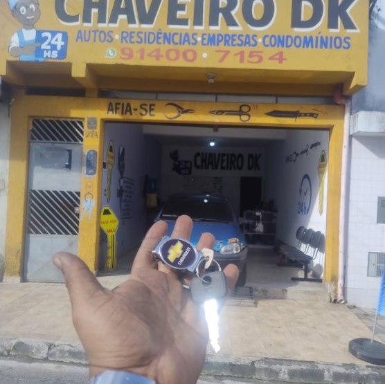 Fachada amarela da loja Chaveiro DK, com placa de atendimento 24 horas, e a mão do chaveiro segurando a chave de um carro
